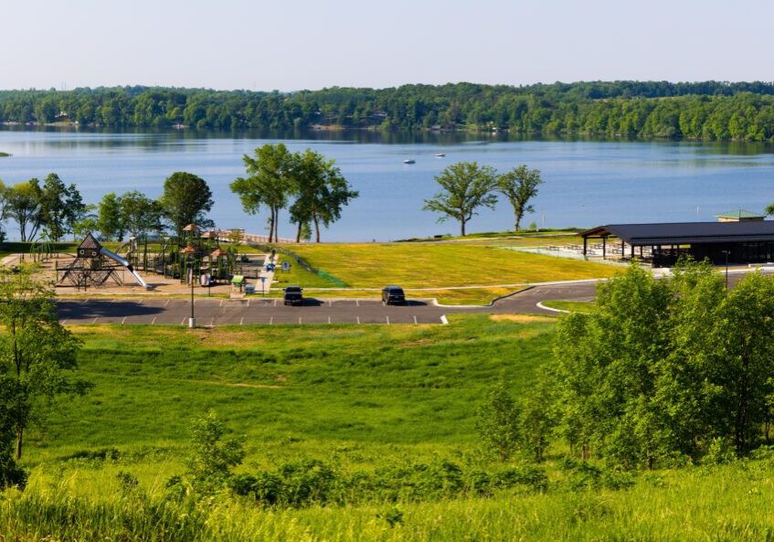 Lake Brophy County Park features a playground, picnic shelter, and parking lot with a few cars, all surrounded by grass and trees. The park overlooks a calm lake with a forested shoreline in the background.