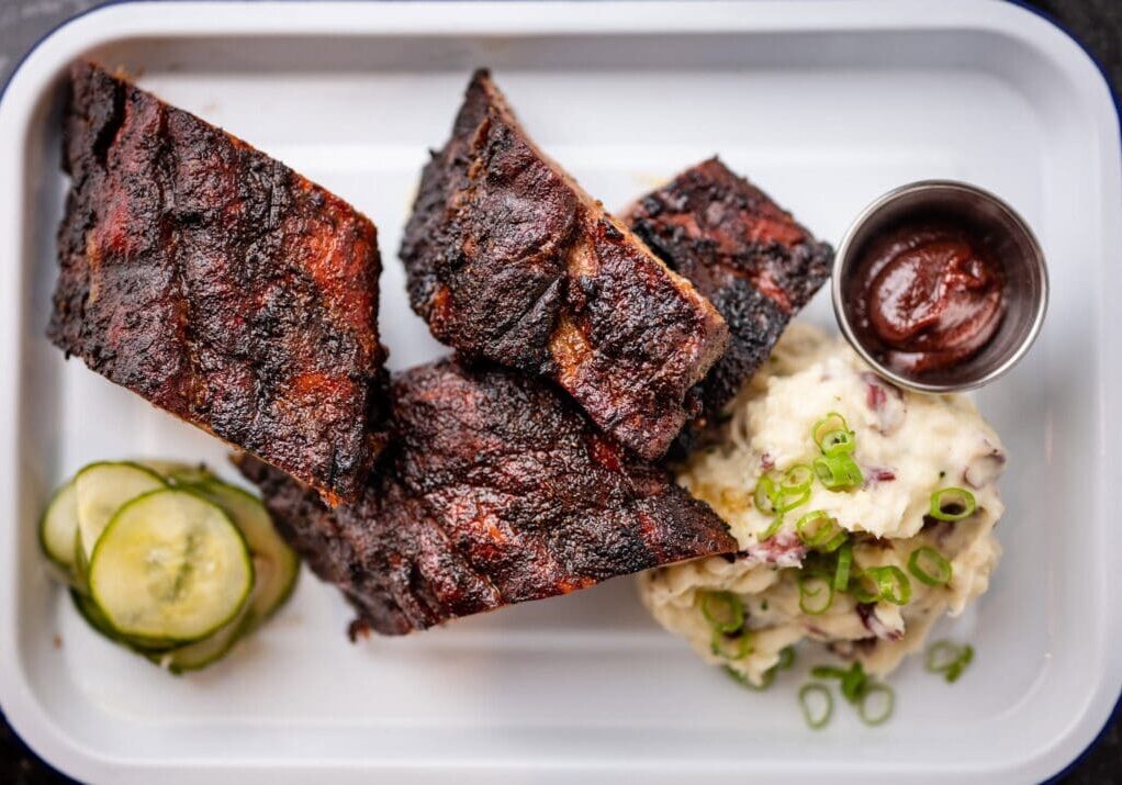 Three pieces of barbecued ribs from Longtrees Woodfire Grill, mashed potatoes topped with chopped green onions, sliced pickles, and a small cup of barbecue sauce served on a white tray.
