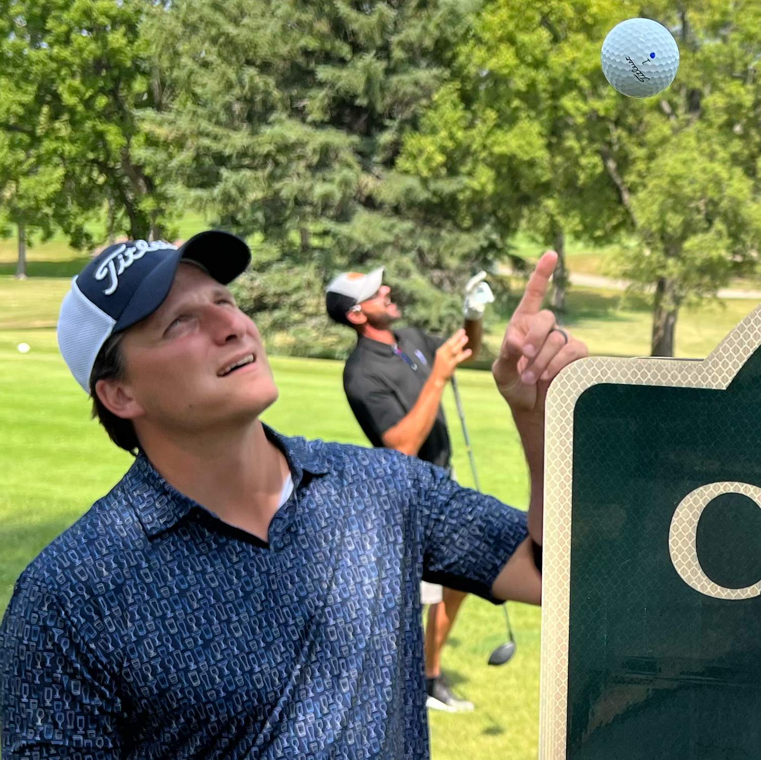 A man in a blue patterned shirt and cap points upwards at a floating golf ball, like a chain of links leading the eye skyward, while another man in the background looks up, both standing on a green golf course with trees.