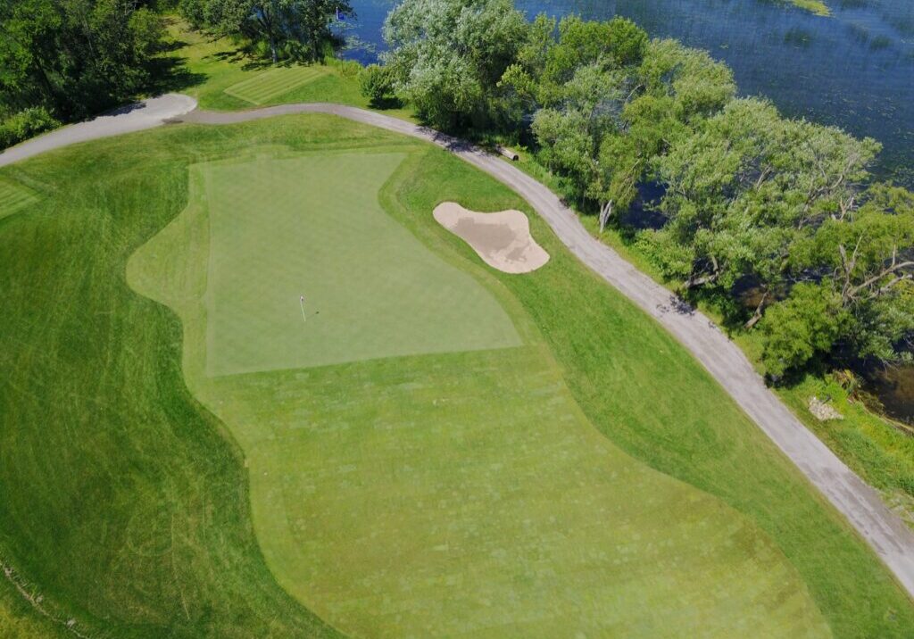 Aerial view of Alexandria Golf Club’s green with a putting hole, surrounding fairway, a sand bunker, a paved path, and trees by a body of water.