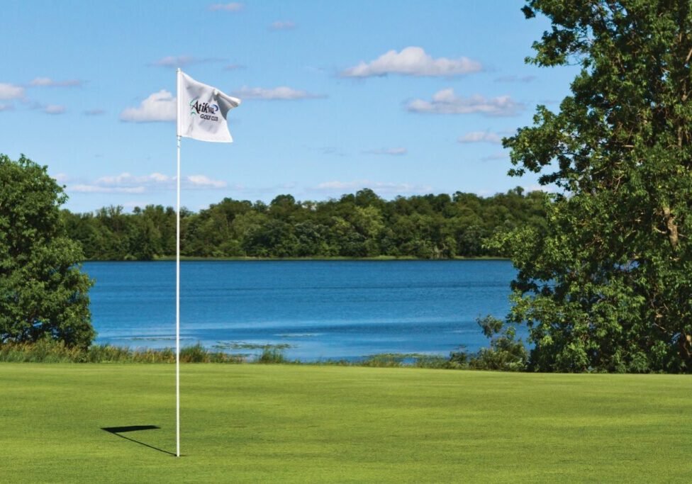 A white golf flag on a green course at Atikwa Golf Course stands near a calm blue lake, surrounded by trees under a partly cloudy sky.