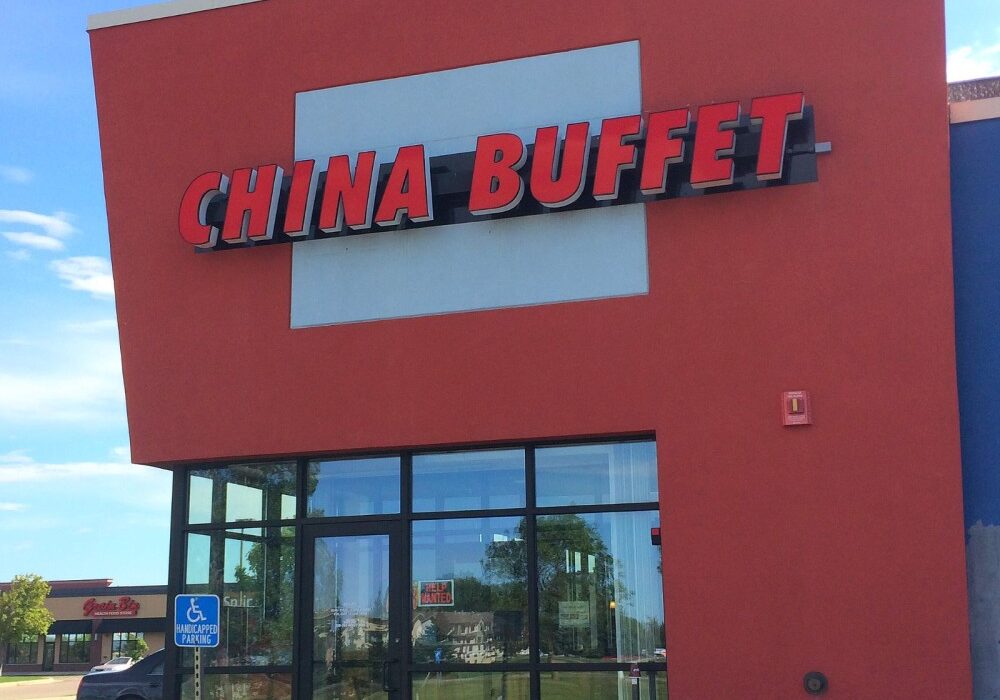 A red building with large windows displays a bright "China Buffet" sign near the entrance, where a blue handicap parking sign stands. The glass reflects nearby trees and other buildings.