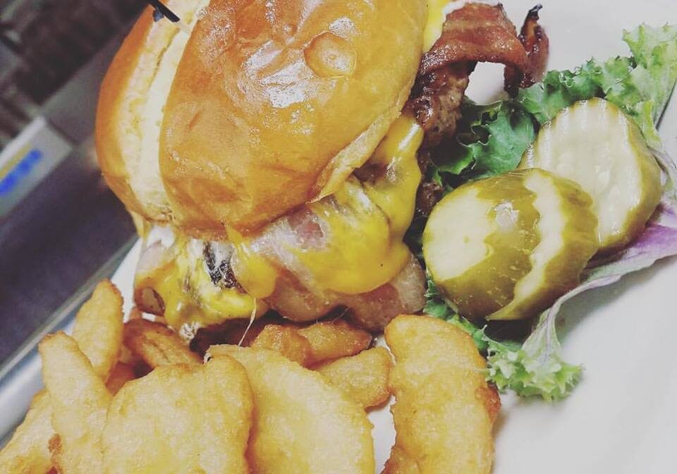 A cheeseburger with bacon on a brioche bun, served with onion rings, sliced pickles, and lettuce on a white plate at Fat Daddy's Bar & Grill.