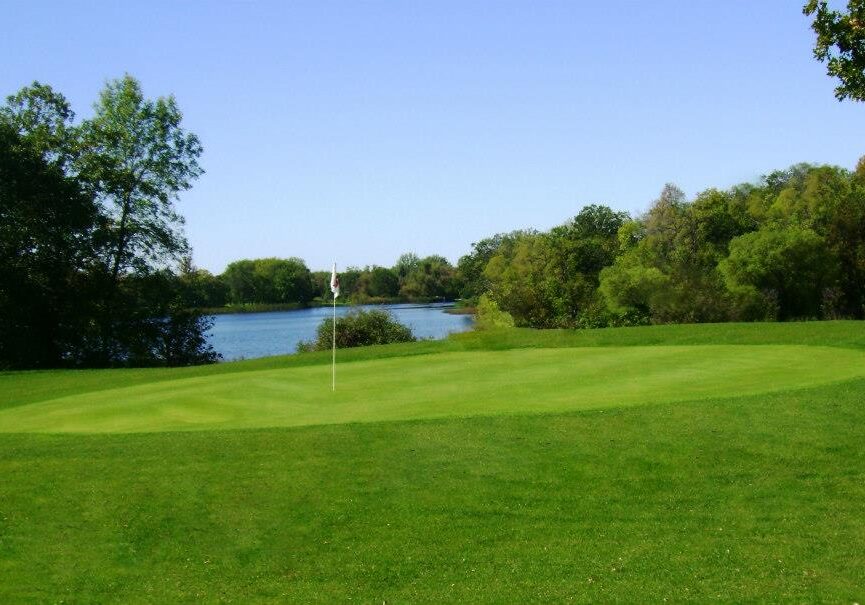 A lush green golf course at Hardwood Hills Golf Course, with a putting green and flagstick in the foreground, surrounded by trees and a calm lake under a clear blue sky.