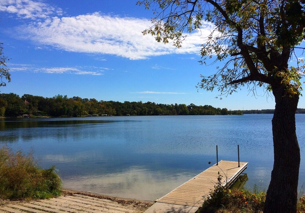 A calm Lake Burgen with clear blue sky, bordered by trees. A wooden dock extends into the water from a gently sloping shore, with a tree in the foreground partially framing the tranquil scene.
