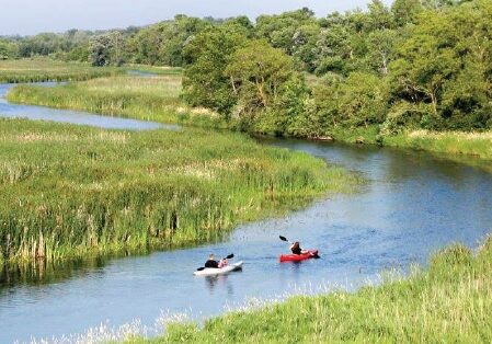 Two people paddle kayaks down the winding Long Prairie River State Water Trail, surrounded by tall green grass and trees under a clear sky.