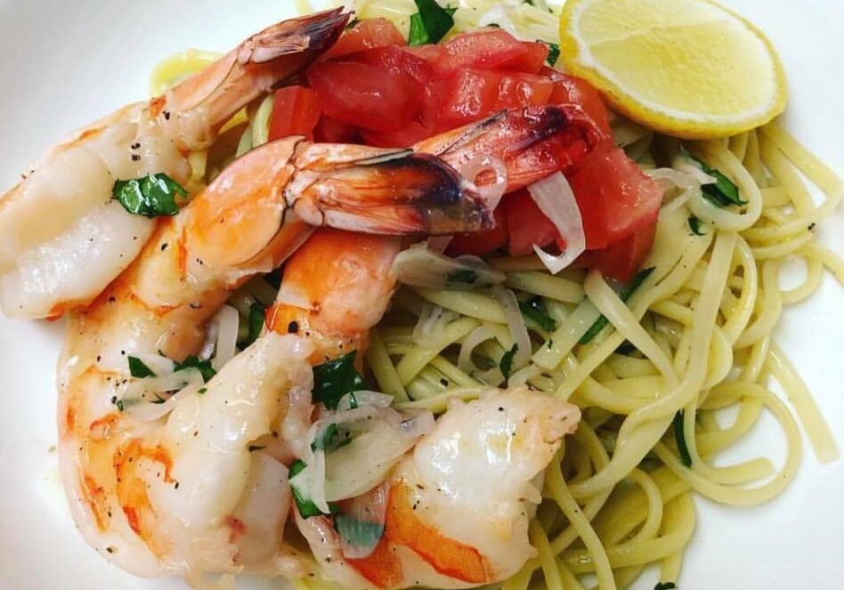 A dish of linguine pasta from Lure Lakebar, topped with grilled shrimp, diced tomatoes, chopped herbs, and onions, served with a lemon wedge on a white plate.