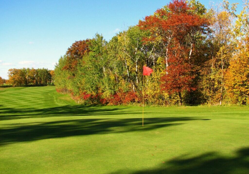 A green golf course at Lake Miltona Golf Club features a red flag on the putting green, bordered by trees with autumn foliage in shades of green, yellow, and orange under a clear blue sky.