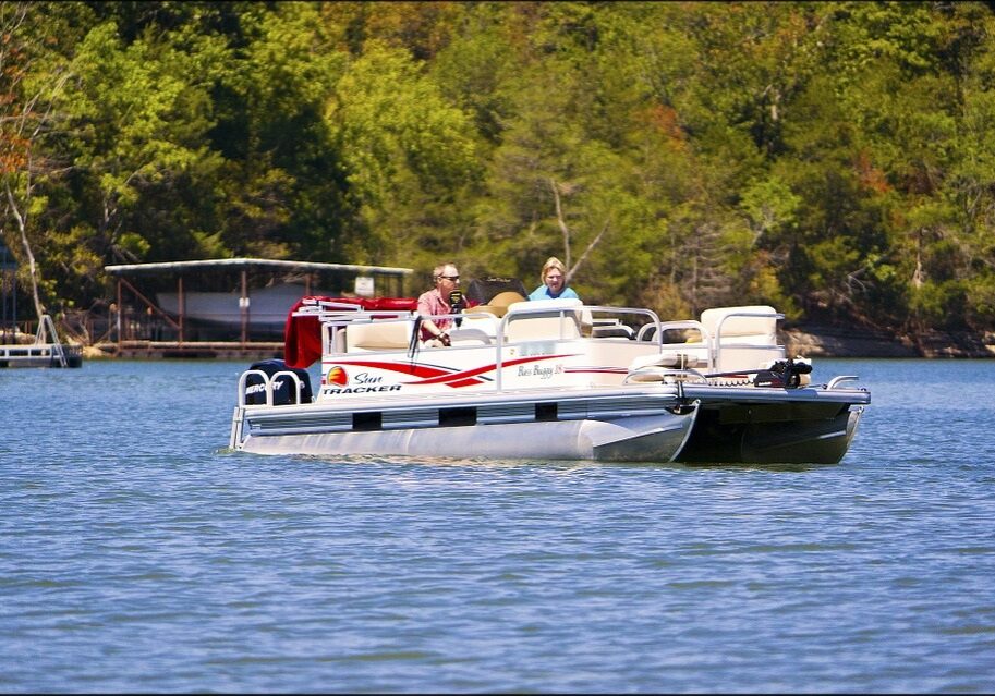 Two people are sitting and relaxing on a Bayside Charters pontoon boat with red and white markings, floating on a calm lake surrounded by trees and greenery on a sunny day.