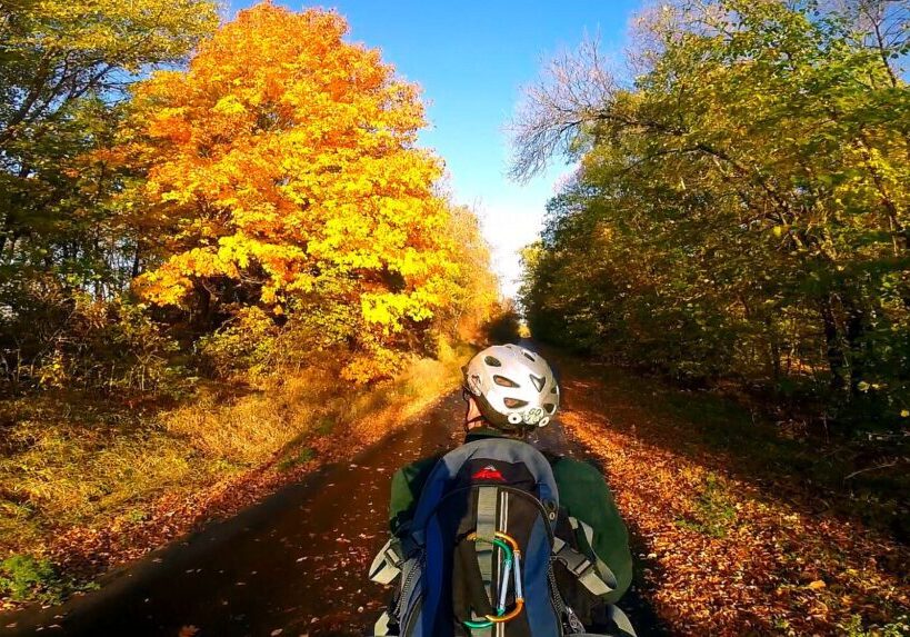 A person biking across Minnesota, wearing a helmet and backpack, cycles along a tree-lined path in autumn, with vibrant yellow and orange foliage under a clear blue sky.